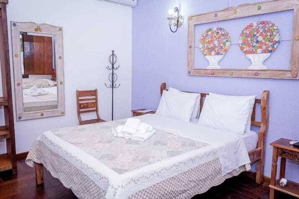 A bedroom with a bed, a wooden chair, a nightstand, a mirror, and a wall-mounted light fixture.