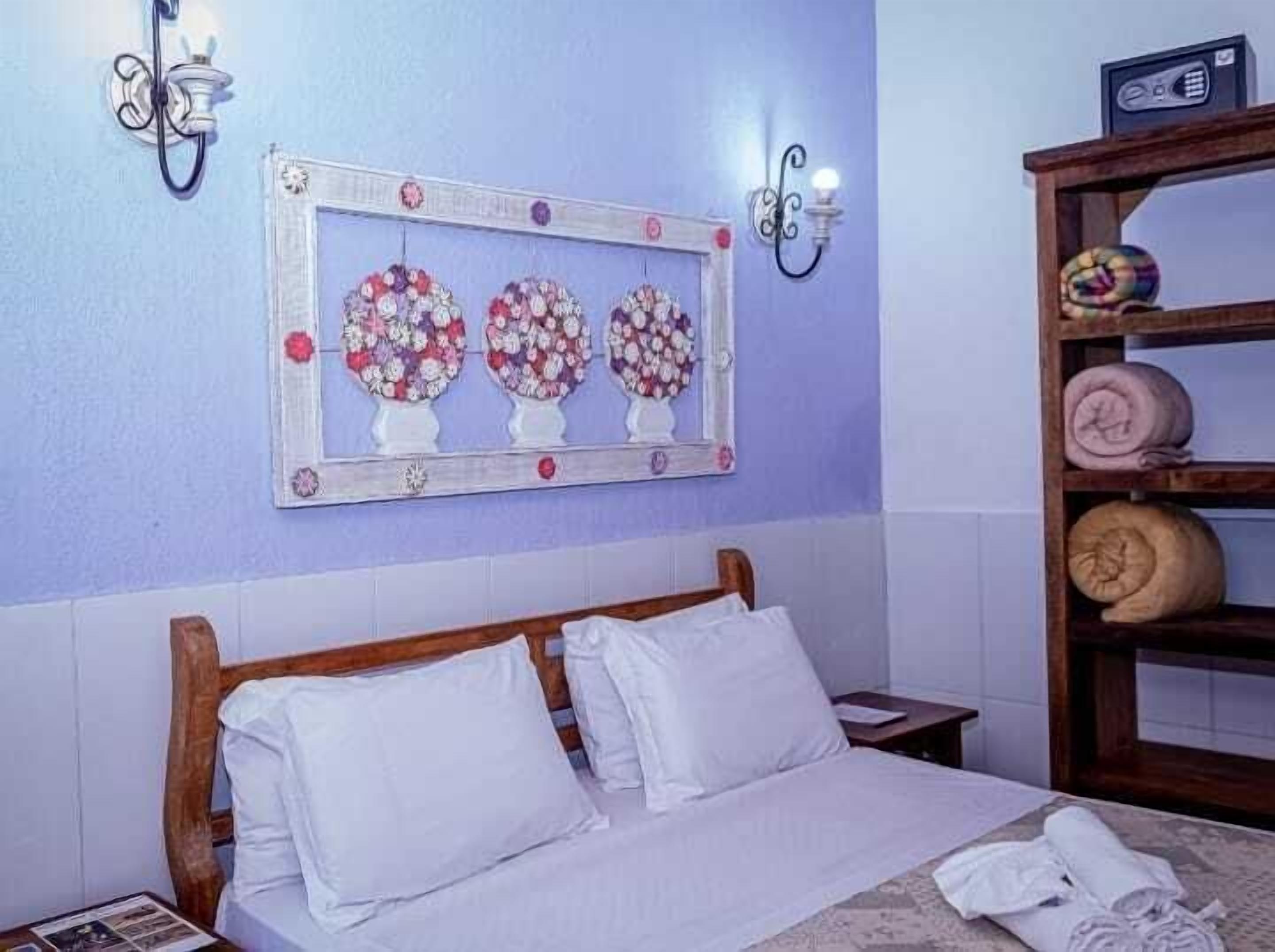 A bedroom with a wooden bed, white bedding, a wall-mounted mirror with floral decorations, and a shelf with rolled towels.
