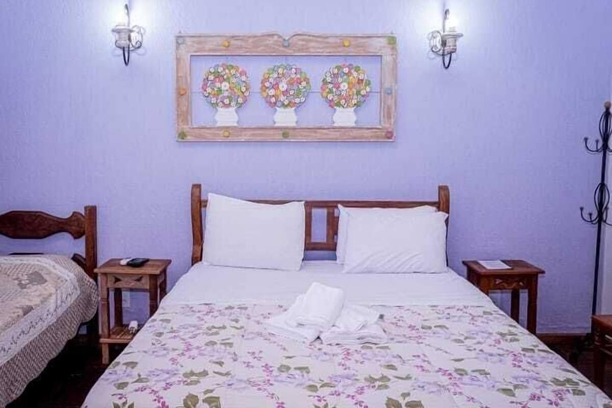 A bedroom with a wooden bed, white bedding, a floral-patterned bedspread, and a wall-mounted picture of three floral arrangements.