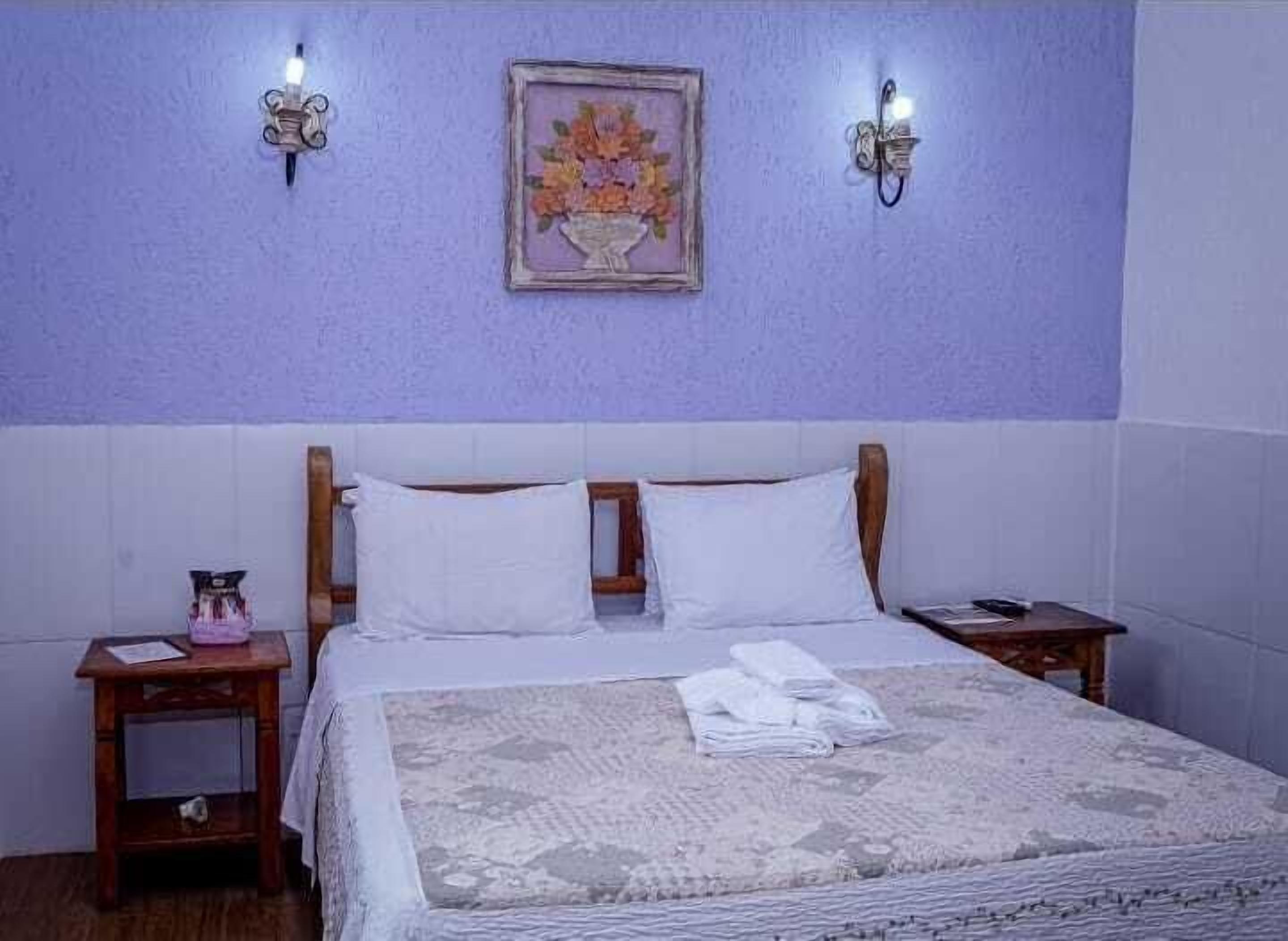 A bedroom with a bed, two bedside tables, a nightstand, a framed floral artwork, and wall-mounted lights.