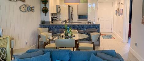 A living room with a blue sofa, a dining area with a table and chairs, and a kitchen in the background.