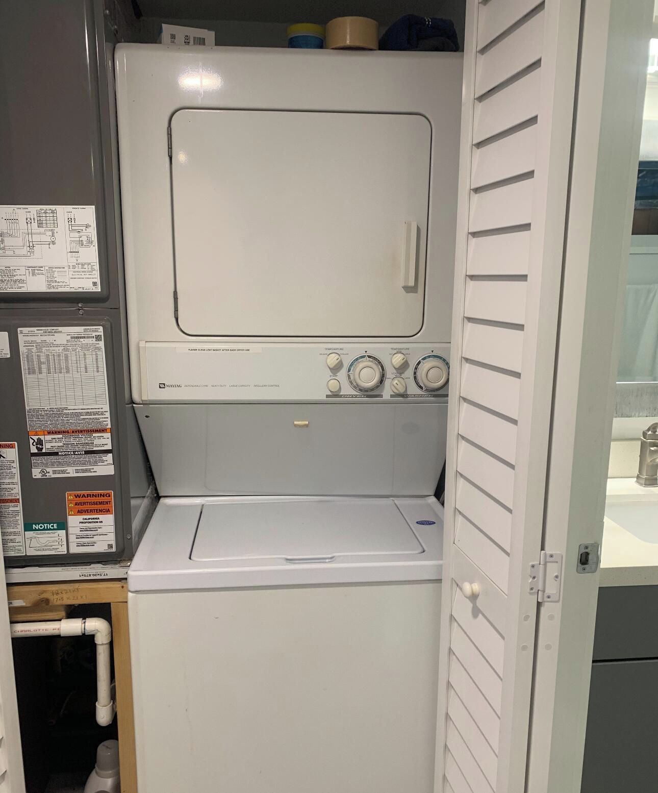A compact, white laundry room with a front-loading washer and dryer. There are warning labels and instructions on the walls, and a small storage area below the dryer.