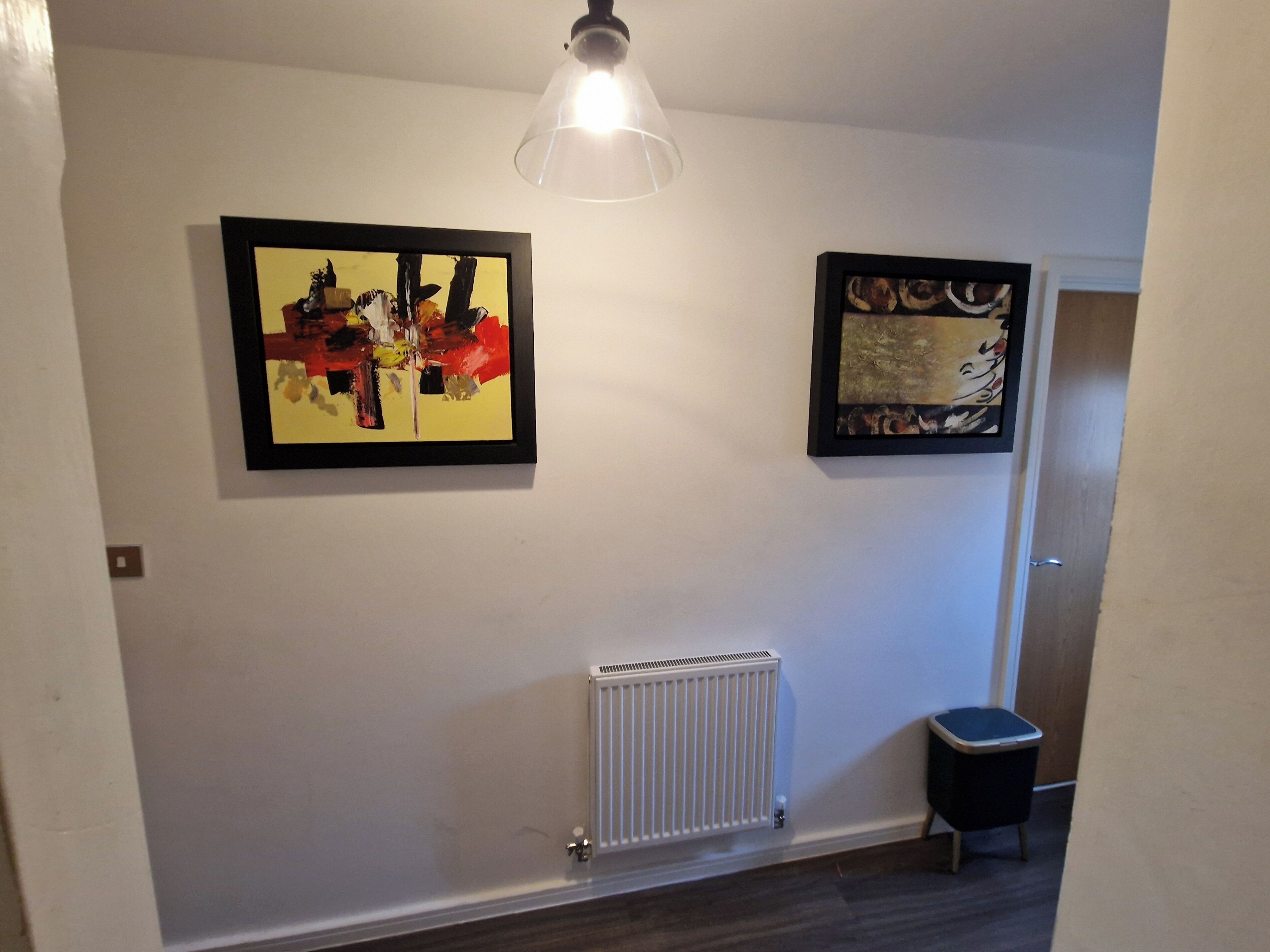 A hallway with two framed abstract paintings, a radiator, and a trash bin.