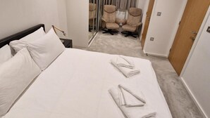 Una cama bien hecha con dos toallas dobladas, una mesita de noche con lámpara, y una silla al fondo.