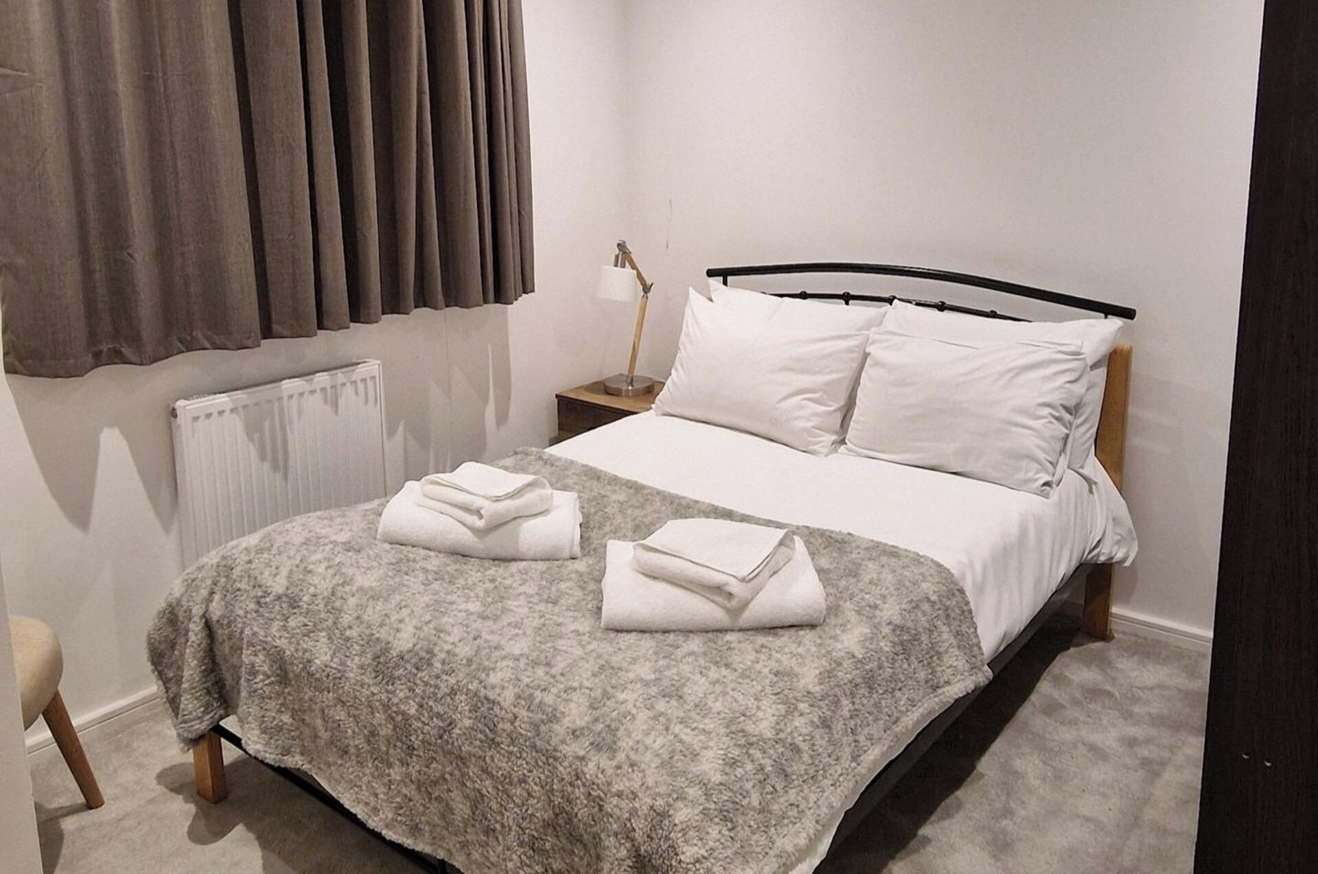 A neatly made bed with white linens and a grey blanket, a bedside table with a lamp, and a radiator on the wall.