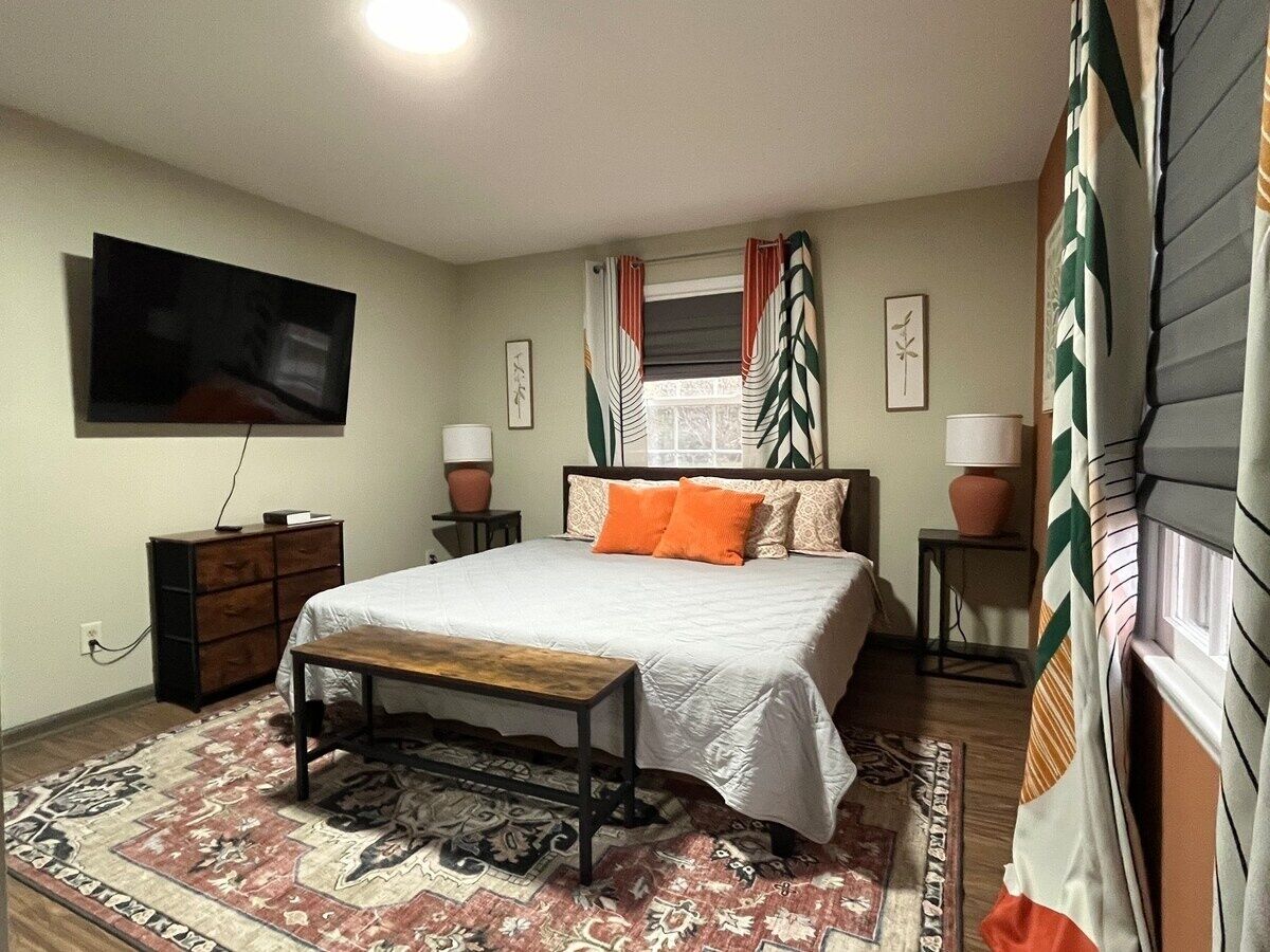 Une chambre à coucher comprenant un lit, un téléviseur fixé au mur, une commode en bois, deux tables de chevet avec des lampes et un tapis à motifs.