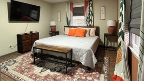 A bedroom with a bed, a TV mounted on the wall, a wooden dresser, two bedside tables with lamps, and a patterned rug. - The Boho Blast of Winston-Salem (Winston-Salem)