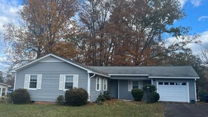 A two-story house with a garage, a front lawn, and mature trees in the background. - The Boho Blast of Winston-Salem (Winston-Salem)