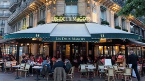 Un café al aire libre con mesas y sillas, un área de estar cubierta y un edificio con un letrero que dice 'LES DEUX MAGOTS'.