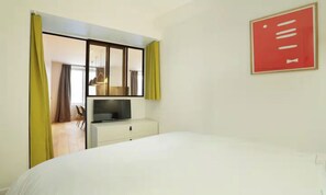 A hotel room with a bed, a TV on a stand, a desk with a chair, and a view of the dining area through a glass door.