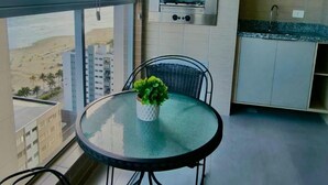 A balcony with a view of a beach and buildings, a round table with a plant, a grill, and a sink. - Residencial Acquamare (Praia Grande)