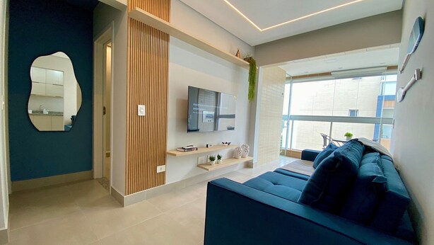A modern living room with a blue sofa, a flat-screen TV, and a large window. - Residencial Acquamare (Praia Grande)