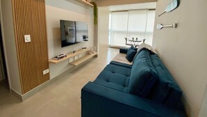 A modern living room with a flat-screen TV, a sofa, a coffee table, and a dining area with a glass partition. - Residencial Acquamare (Praia Grande)