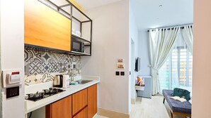 A compact kitchen with a gas stove, a coffee machine, and a microwave. There is a small dining area with a sofa and a table, and a window with curtains. - Casa- Grey Apartments (Abuja)