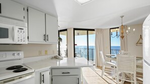 A modern kitchen with white cabinetry, a microwave, and a stove. There is a dining area with a table and chairs, and a balcony with a view of the sea. - Phoenix IV 4112 (Orange Beach)