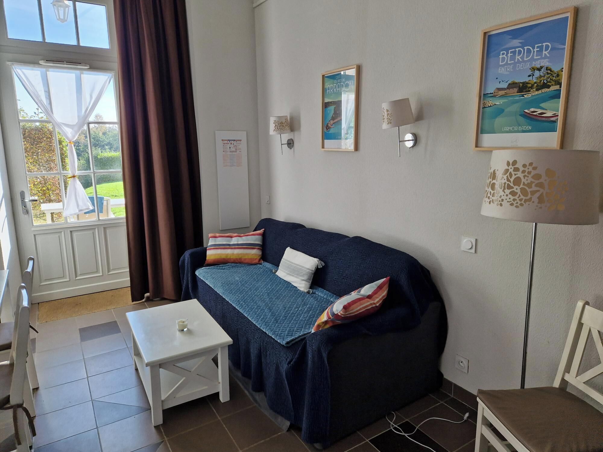 Una habitación acogedora con un sofá azul, una mesa blanca y una ventana con cortinas. Hay lámparas fijadas a la pared y un cuadro enmarcado.