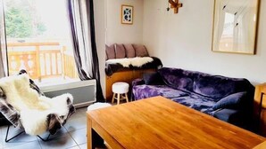 A living room with a wooden coffee table, a purple sofa, a white furry chair, and a small stool. There is a window with curtains, a wooden deck outside, and a framed picture on the wall.