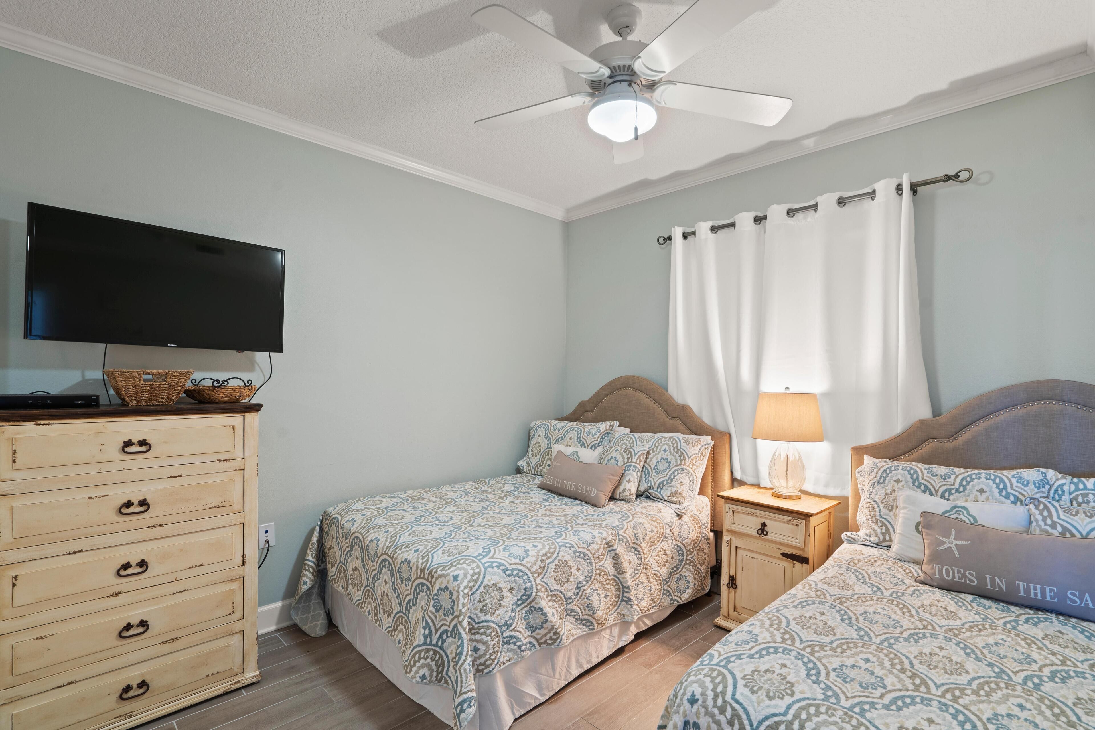 Une chambre avec deux lits, une commode, un téléviseur et un ventilateur de plafond.