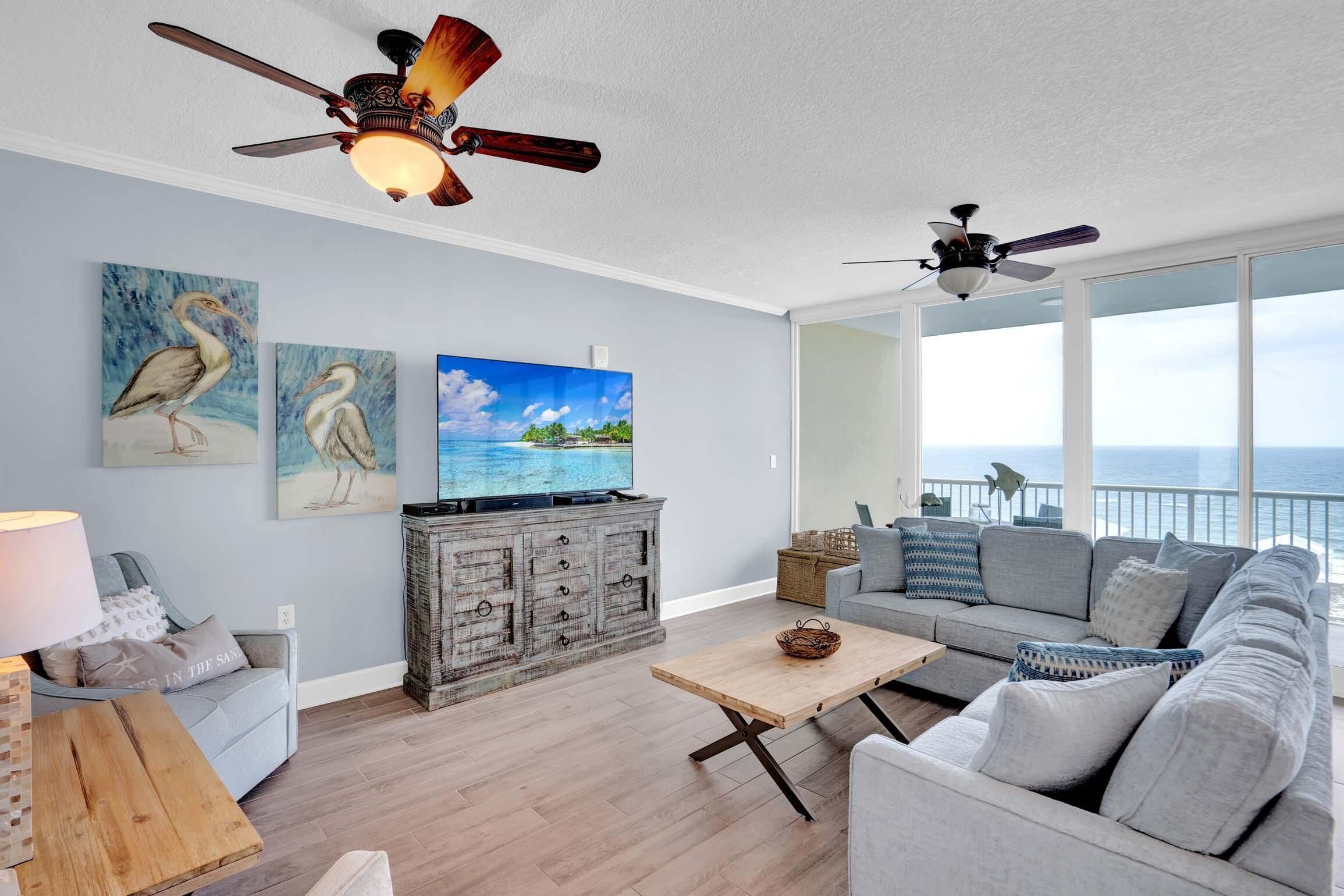 A spacious living room with a large window overlooking the ocean, a ceiling fan, and a television on a wooden cabinet.