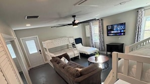 A spacious room with a high ceiling, a ceiling fan, a flat-screen TV mounted on the wall, a brown sofa, a white crib, and a small round table with a plant. - The Bunkhouse at Origins Ranch (Plant City)