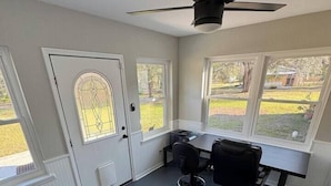 A room with a ceiling fan, a desk with two chairs, a black leather bench, and large windows with a view of the outdoors. - The Bunkhouse at Origins Ranch (Plant City)
