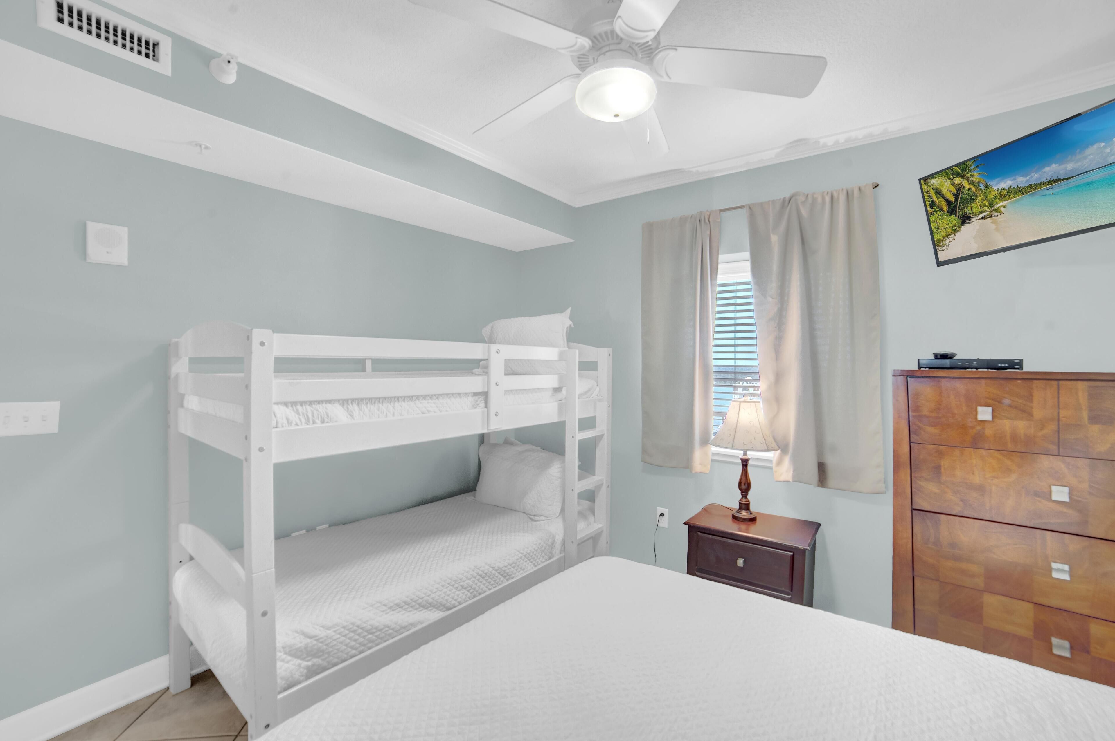 Un dormitorio con literas, ventilador de techo, cómoda de madera y ventana con cortinas.