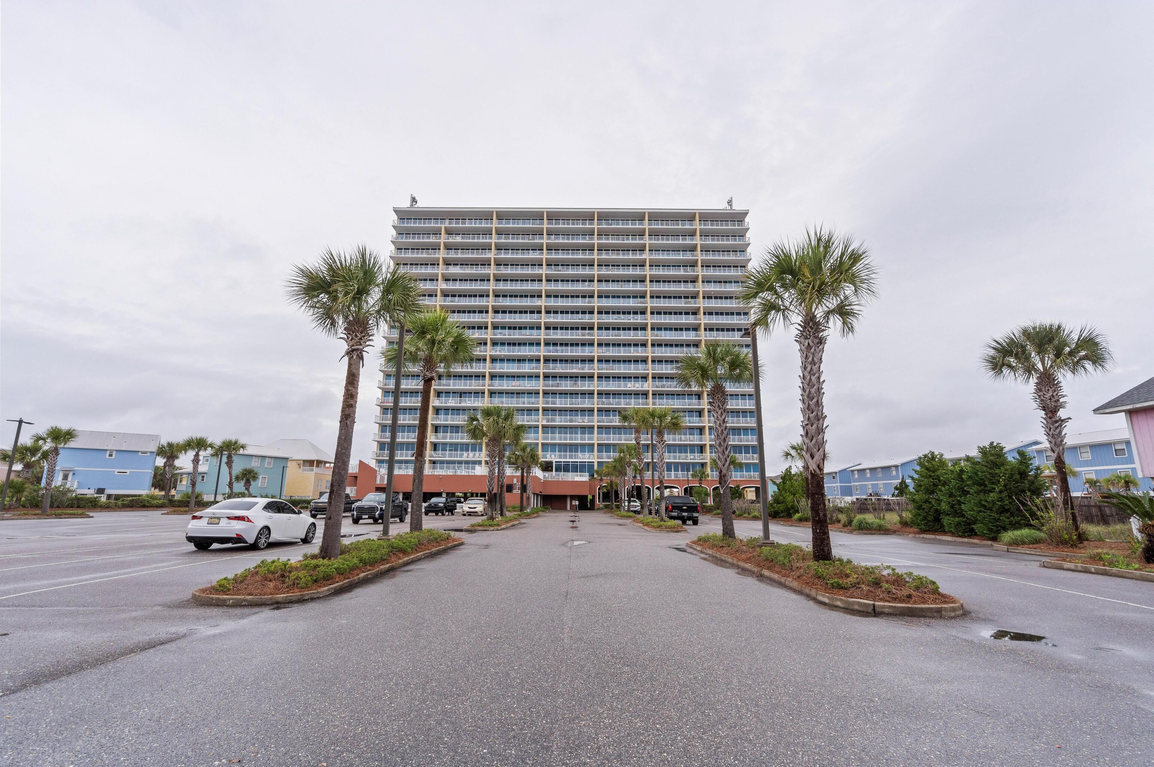 A multi-story hotel with a central entrance, surrounded by palm trees and a parking lot.