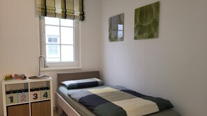 A bedroom with a single bed, a window with blinds, a bedside table with a lamp, and a storage unit with numbered drawers.