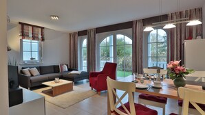 A modern living room with a sofa, red chairs, a dining table, and large windows. - This enchanting, modern and tastefully furnished 3-room apartment with a covered terrace and a view of the countryside invites you to linger. (Ostseebad Nienhagen)