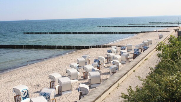 A beach with rows of white beach chairs and umbrellas, a wooden walkway, and a pier extending into the water. - This enchanting, modern and tastefully furnished 3-room apartment with a covered terrace and a view of the countryside invites you to linger. (Ostseebad Nienhagen)
