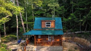 A small cabin with a green roof, wooden exterior, and a porch with a bench.