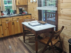 Une cuisine rustique aménagée dans une cabane en bois, avec une table à manger, des chaises, une fenêtre avec des stores et une vue sur l’extérieur.