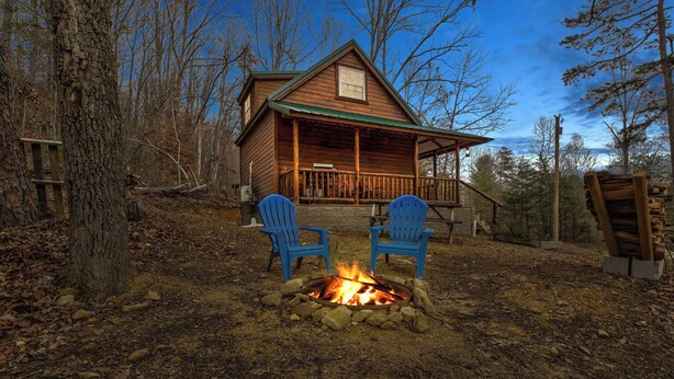 A cabin with a fire pit and two blue chairs in front.