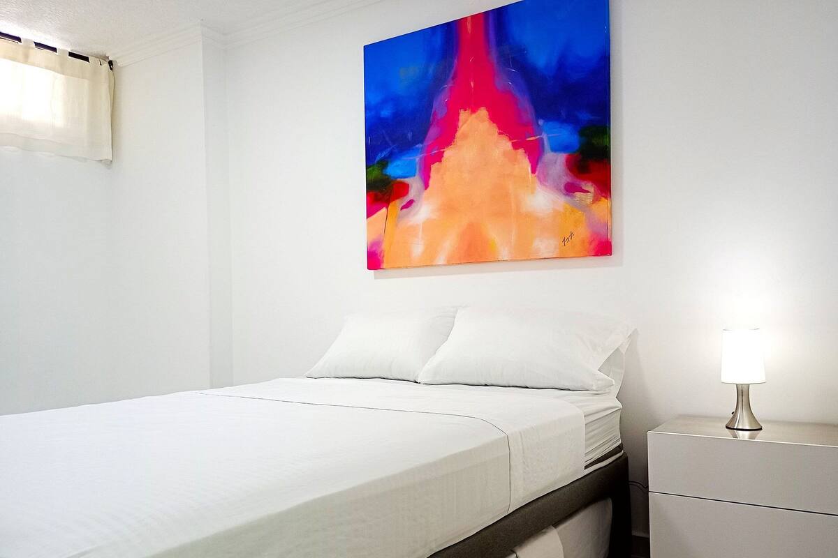 A bed with white linens, a nightstand with a lamp, and a colorful abstract painting on the wall.