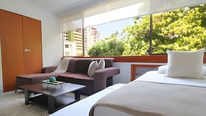 Quarto de hotel moderno com sofá, uma mesa pequena, uma cama e uma janela grande com vista para edifícios e árvores.
