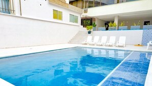 Piscina com água azul cristalina, rodeada por espreguiçadeiras brancas e plantas em vasos.