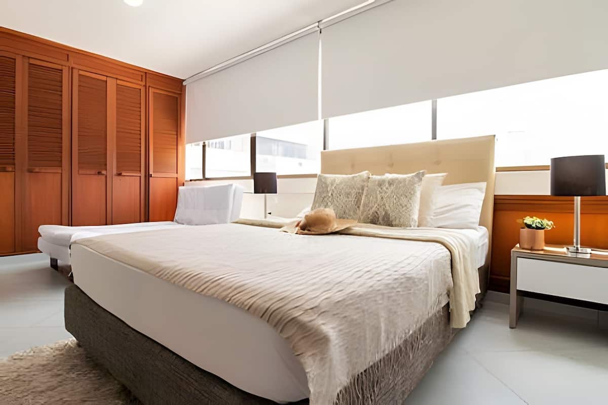 A bedroom with a large bed, wooden wardrobe, bedside tables, and a window with blinds.