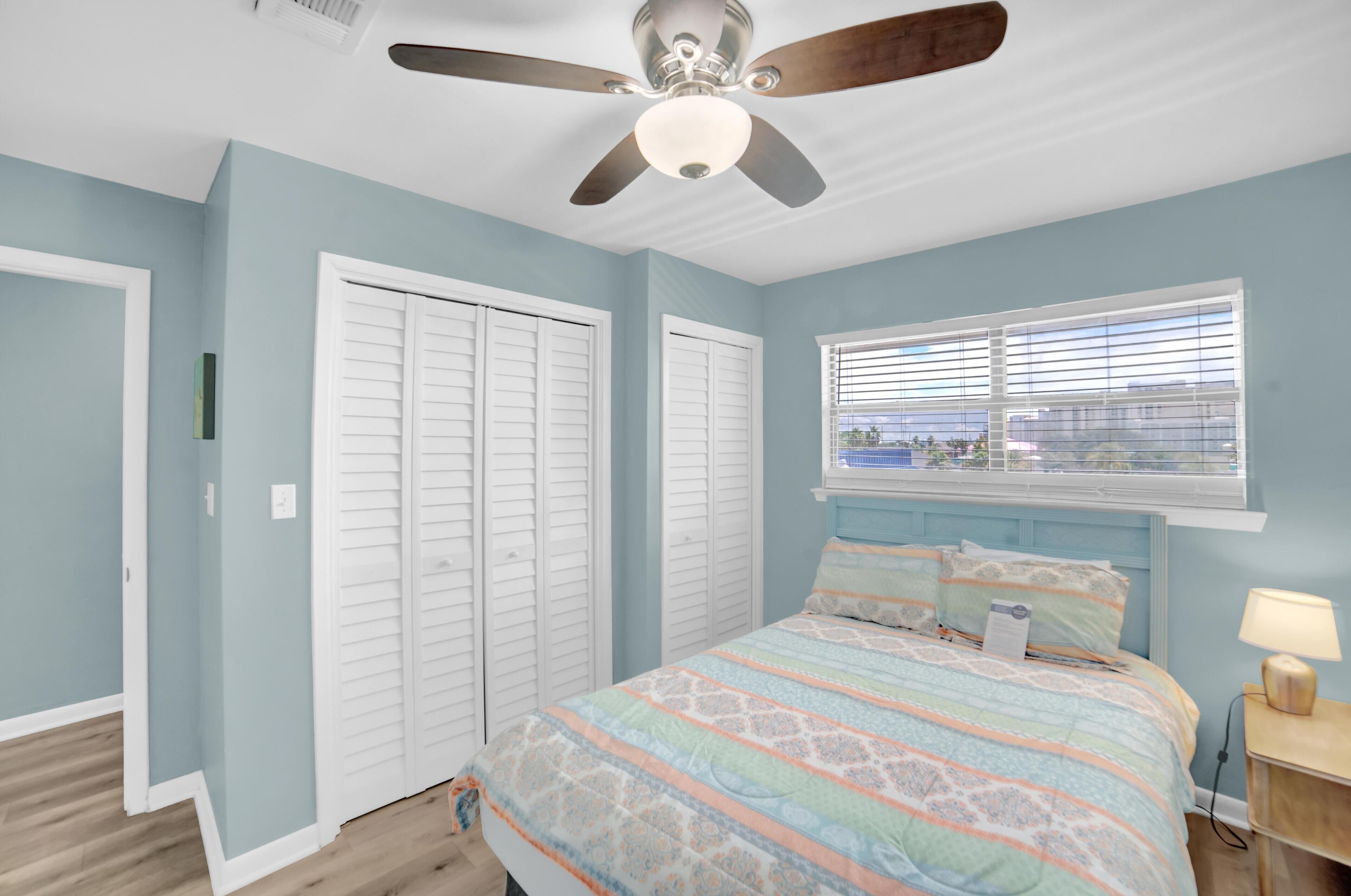 Une chambre à coucher avec un lit, un ventilateur de plafond, une fenêtre avec des stores et une table de chevet avec une lampe.