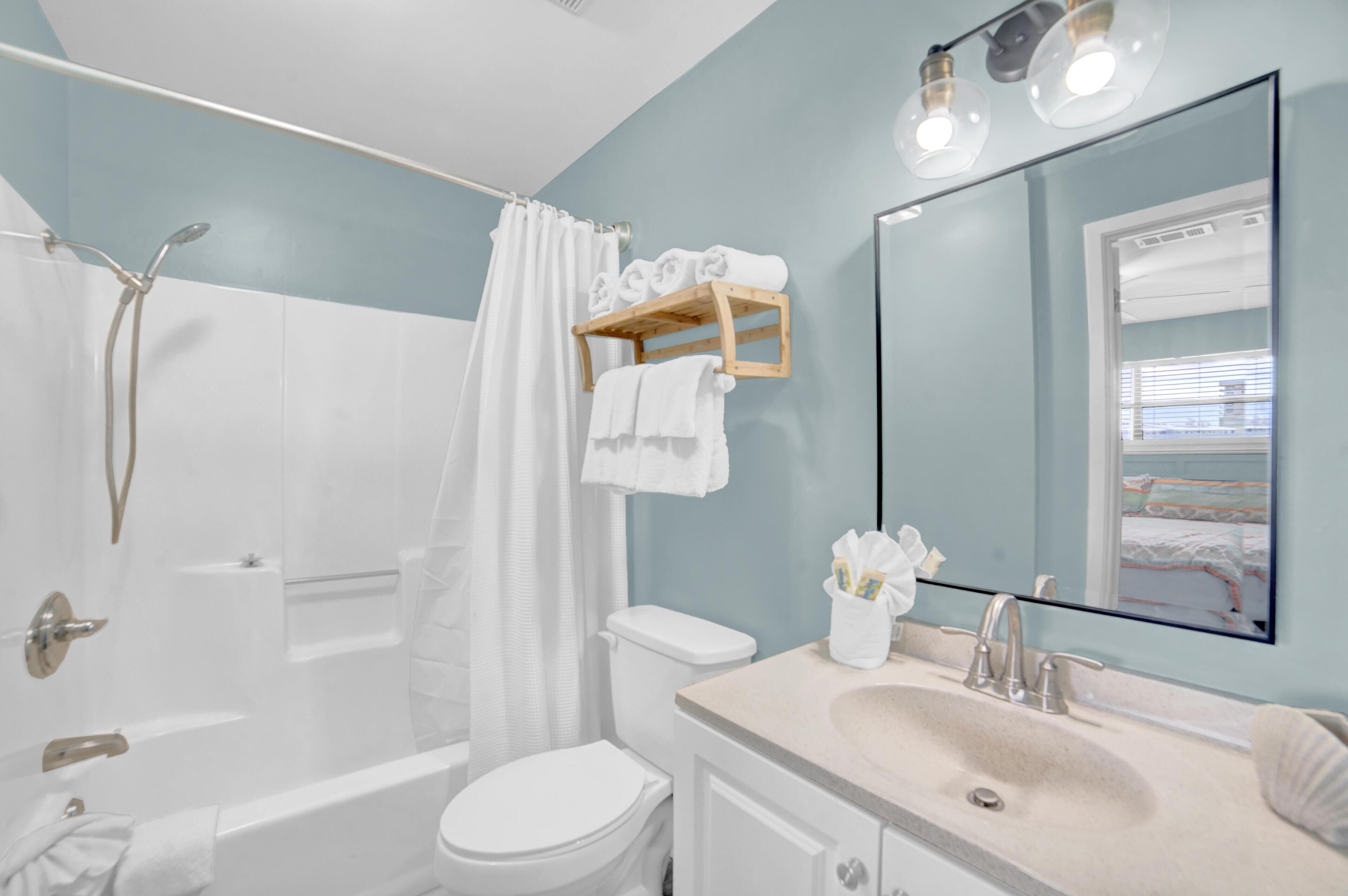 A modern bathroom with a bathtub, toilet, and sink. There is a shower curtain, a towel rack, and a mirror with a view of the outside.