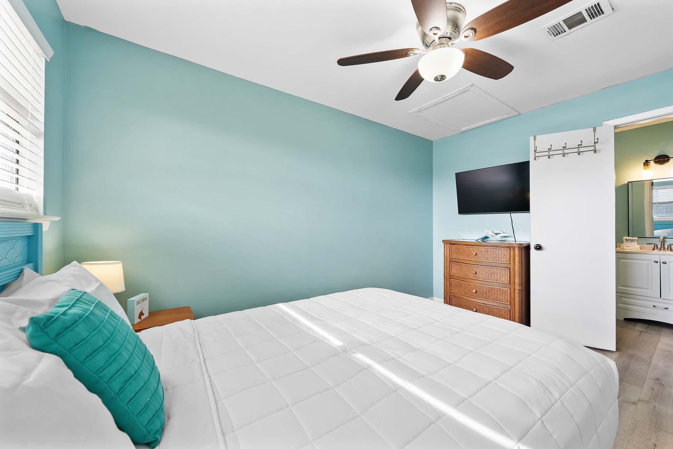 A bedroom with a bed, a ceiling fan, a TV, and a bathroom visible through an open door.