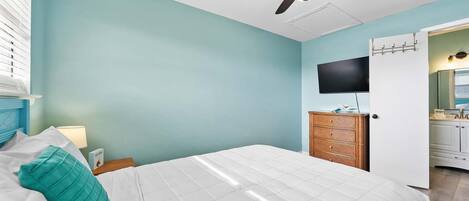 A bedroom with a bed, a ceiling fan, a TV, and a bathroom visible through an open door.