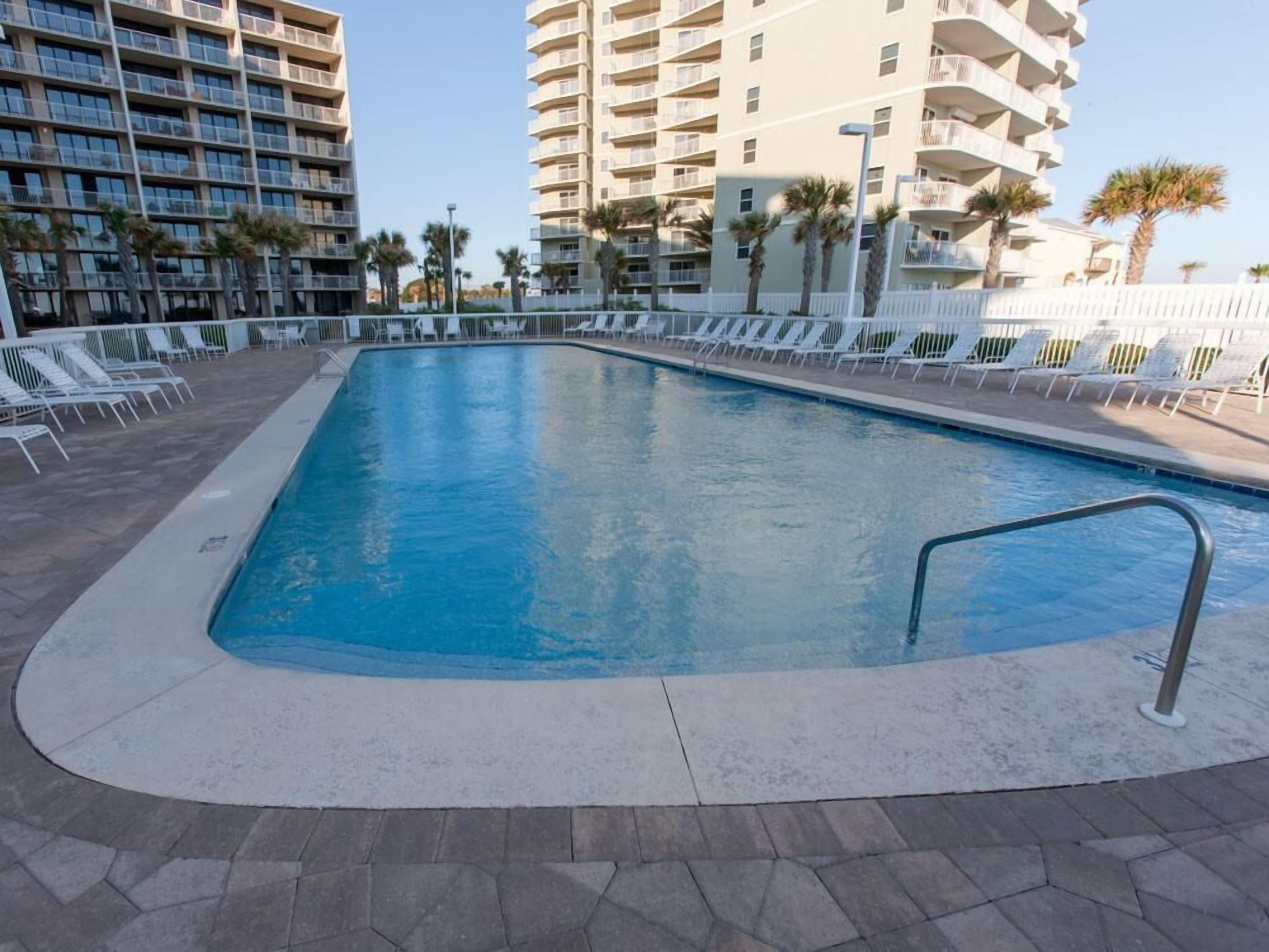 A swimming pool with a tiled deck, surrounded by white lounge chairs and palm trees.