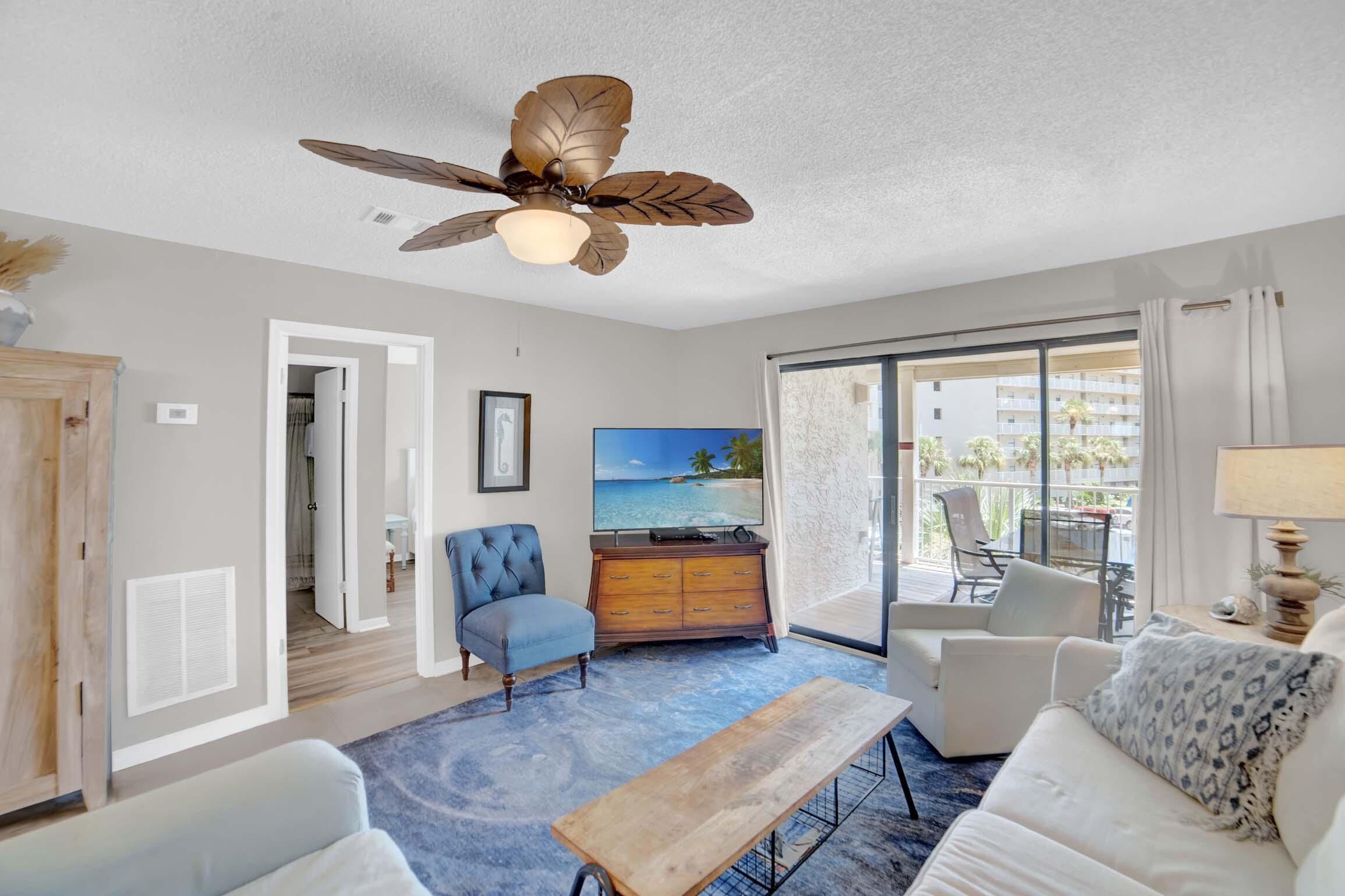 A spacious living room with a ceiling fan, a large TV, a wooden coffee table, and a view of the outdoors through a sliding glass door.