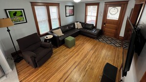 A living room with a sectional sofa, a small table, and a green ottoman. There are two windows with blinds, a ceiling light, and a door with a glass panel.