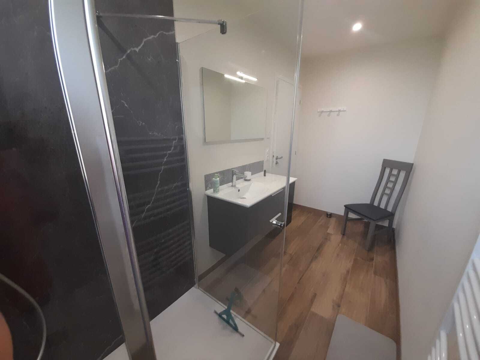 Une salle de bain moderne avec une douche à parois de verre, un lavabo blanc et une chaise en bois.