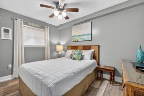 A bedroom with a bed, a nightstand, a lamp, a ceiling fan, a window with curtains, a wall-mounted artwork, and a wooden side table. - Seaside Beach & Racquet 3305 (Orange Beach)