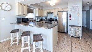 A kitchen with a central island, bar stools, a microwave, stove, refrigerator, and a clock on the wall. - Seaside Beach & Racquet 3401 (Orange Beach)