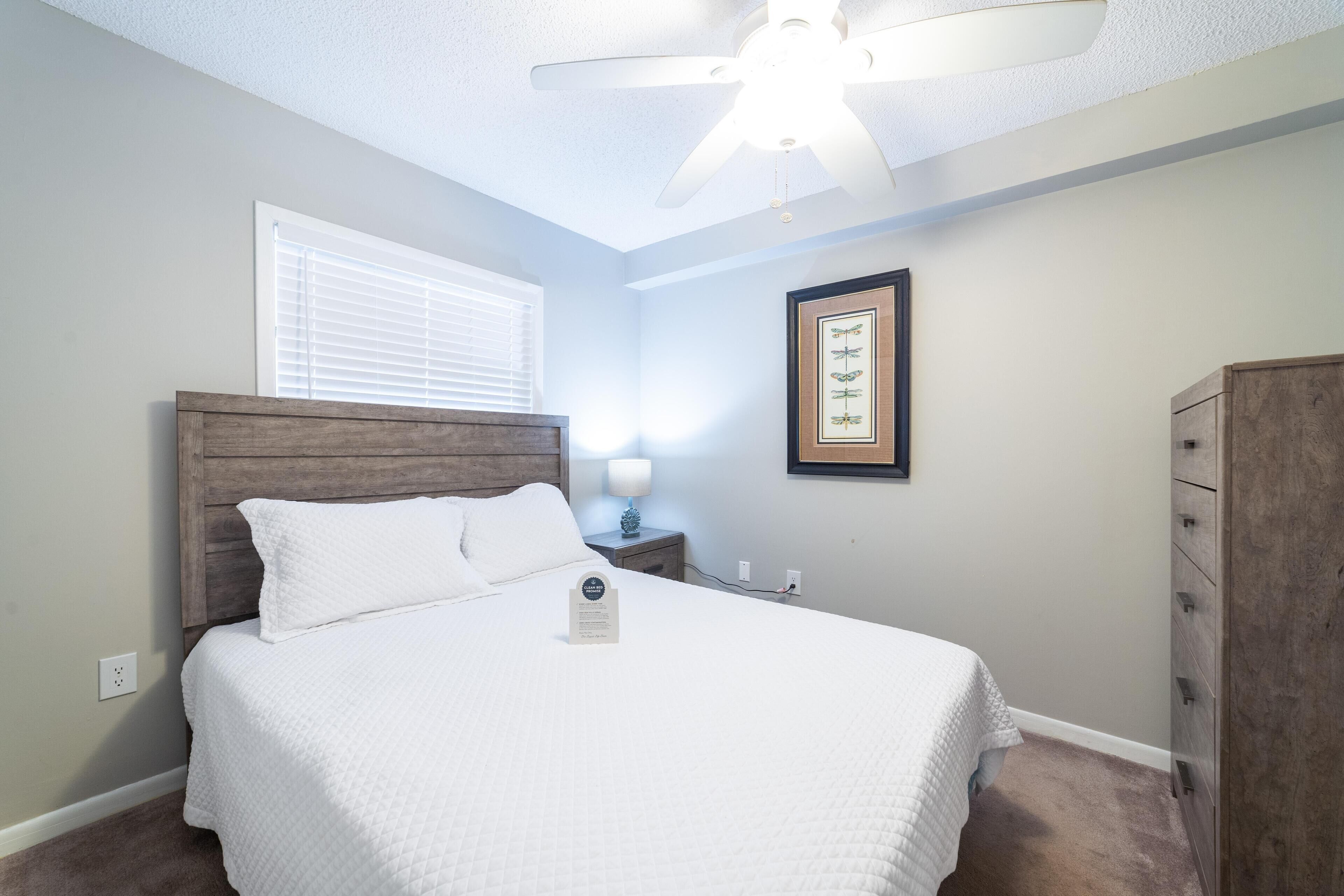 A bedroom with a bed, a nightstand, a ceiling fan, a window with blinds, a framed picture on the wall, and a wooden dresser.