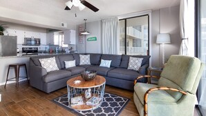 A modern living room with a grey sectional sofa, a round coffee table, and a bamboo chair. A ceiling fan and a kitchen are visible in the background. - Seaside Beach & Racquet 4412 (Orange Beach)
