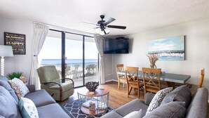A modern living room with a large window, a ceiling fan, a dining area, and a TV mounted on the wall. - Seaside Beach & Racquet 4412 (Orange Beach)